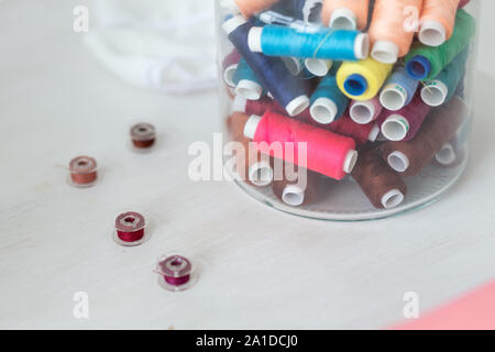 Close up multicolored threads and bobbins lie on a table in a transparent glass jar on the seamstress's desktop. The concept of unique designer clothe Stock Photo