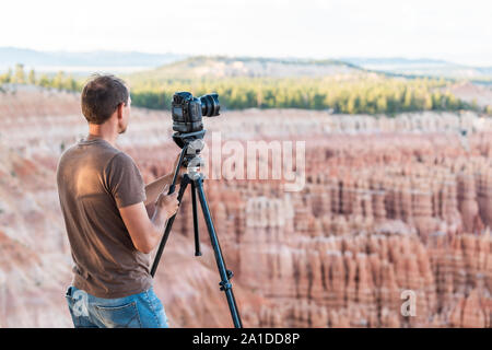 Man taking pictures of view from Bryce Point overlook of hoodoos rock formations in Bryce Canyon National Park at sunset with tripod and camera Stock Photo