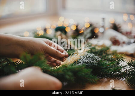 Hands holding herbs and fir branches, pine cones, thread, berries, golden lights on wooden table. Christmas wreath workshop. Authentic stylish still l Stock Photo