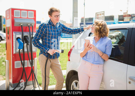 Man and woman are standing near the electric car. The car is charging at the charging station for electric vehicles. Stock Photo