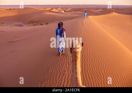 Two nomadic men dressed in traditional clothing lead a camel through sand dunes toward a tented camp in the Sahara Desert, Morocco. Stock Photo