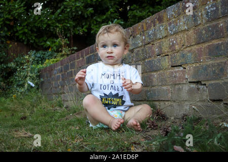 A muddy small caucasian boy (age 11 months) with blue eyes wearing a grubby white t-shirt top sits in the garden by a brick wall looking worried Stock Photo