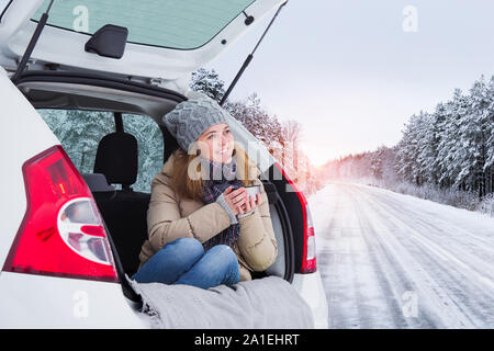 Young woman traveler sits in the trunk of the car and holds a cup of hot tea in hands. Stock Photo