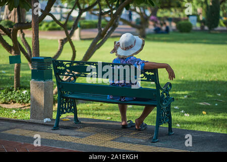 A Thai woman wearing a hat relaxes on a bench in Santichaiprakarn Park, in Banglamphoo area, Bangkok, Thailand Stock Photo