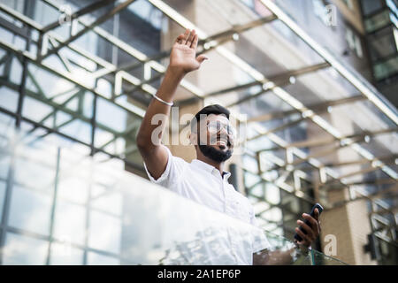 Serious indian businessman in eye glasses looking on copy space while standing against glass skyscraper waving hello to someone. Stock Photo