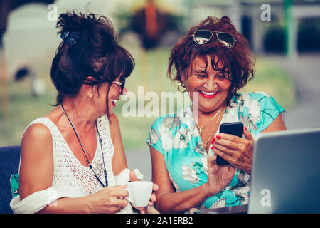 Cheerful senior ladies using a smart phone and talking while sitting outdoors in a cafe Stock Photo