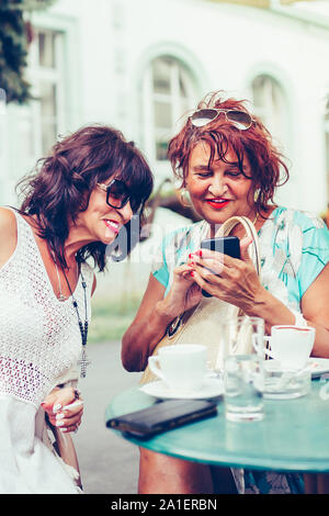 Cheerful senior ladies using a smart phone, laughing and talking while sitting outdoors in a cafe Stock Photo