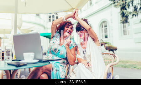 Happy senior women are giving high five, celebrating success while looking each other sitting in the table in a outdoor cafe. Stock Photo