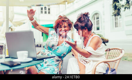 Two happy senior women using laptop and pointing at screen surprised while sitting in outdoor cafe Stock Photo