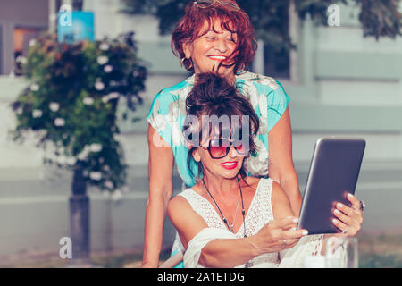 Two happy senior women using tablet and watching media content online while sitting in a outdoor cafe. Stock Photo