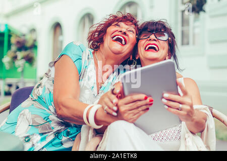 Two excited senior women sitting in outdoor cafe laughing and watching social media and funny videos on a tablet. Stock Photo