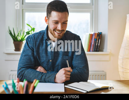 Single happy young bearded man with pen in hand looking down at notebook on his desk while seated in front of bright window Stock Photo