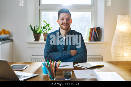 Confident smiling bearded man with folded arms in casual clothes sitting at desk in front of bright window Stock Photo