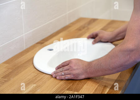 Closeup of hands of professional plumber worker installs white oval ceramic sink on wooden tabletop in bathroom with beige tile Stock Photo