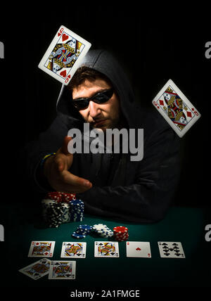 Man dressed in hood and sunglasses at a table playing poker shows his cards, throwing them at the camera, showing that he has achieved a queens poker. Stock Photo