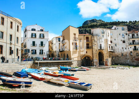 Cefalu, Sicily, Italy - Apr 7, 2019: Beautiful old harbor in small Sicilian city. Boats on a sandy beach. Traditional houses, mountain in the background. People on the promenade. Sunny day. Stock Photo