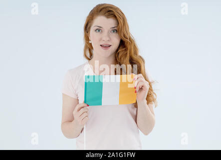 Red haired young woman with the flag of Ireland looking at the camera while standing on a light background. Stock Photo