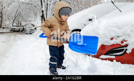Close up shot of smiling charming boy in beige jacket and grey hat helping to clean up the snow covered red car after snow storm using big blue shovel Stock Photo