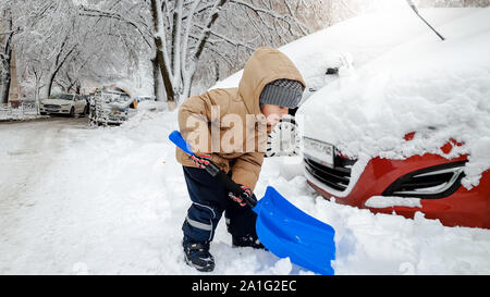 Closeup photo of smiling charming boy in jacket and grey hat helping to clean up the snow covered red car after snow storm using big blue shovel Stock Photo