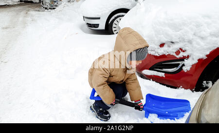 Closeup shot of smiling charming boy in jacket and hat helping to clean up the snow covered car after blizzard using big blue shovel Stock Photo