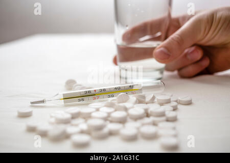 In medical thermometer temperature 37 degrees, and in the background are on the table white pills and hand reaches for a glass of water Stock Photo