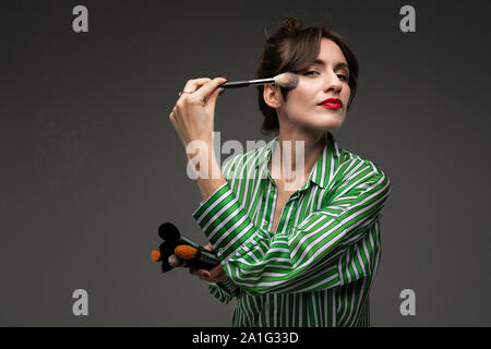 Portrait of young woman putting on makeup on cheeks in green and white striped blouse with makeup brushes in her hands isolated Stock Photo