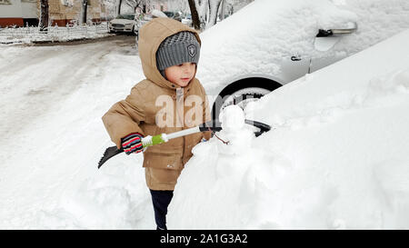 Close up photo of cute smiling boy in warm gloves hat and jacket helping to clean up snow covered parents red car after blizzard using big brush . In Stock Photo