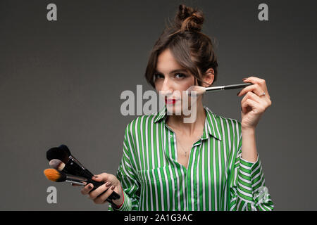 Portrait of young woman putting on makeup in green and white striped blouse with makeup brushes in her hands isolated Stock Photo