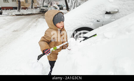 Close up portrait of charming smiling boy in gloves hat and jacket helping to clean up snow covered parents car after blizzard using big brush . In Stock Photo
