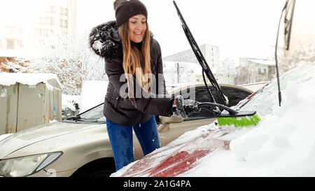 Close up shot of young smiling blond woman in brown jacket hat gloves and blue jeans trying to clean up snow covered red auto by brush after snow Stock Photo