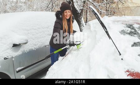 Closeup shot of smiling woman in jacket gloves and jeans trying to clean up snow covered red auto by brush after snow storm. Scraping the windshield Stock Photo