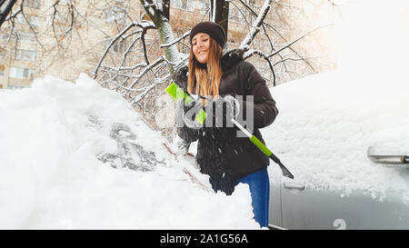 Closeup photo of happy girl in jacket gloves and jeans trying to clean up snow covered red auto by green brush after snow storm. Scraping the Stock Photo