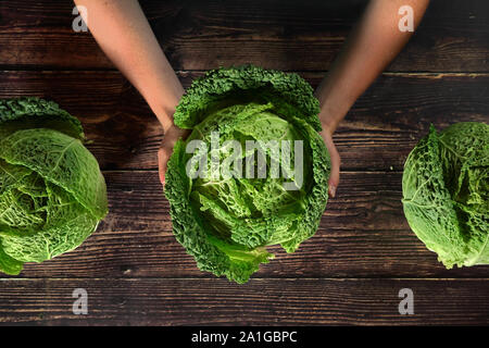 Top down view, hands holding savoy cabbage head over dark wooden board, two other vegetables next to it Stock Photo