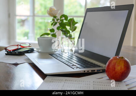Laptop with empty screen, papers, coffee cup, glasses and a fresh apple on a home office workspace near the window, selected focus, narrow depth of fi Stock Photo