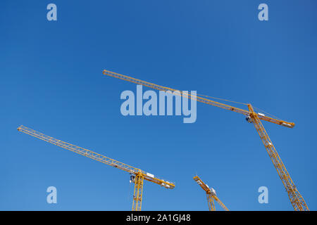 Low angle view group of three yellow tower cranes work over building construction site against clear blue sky. Stock Photo