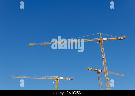 Low angle view group of three yellow tower cranes work over building construction site against clear blue sky. Stock Photo