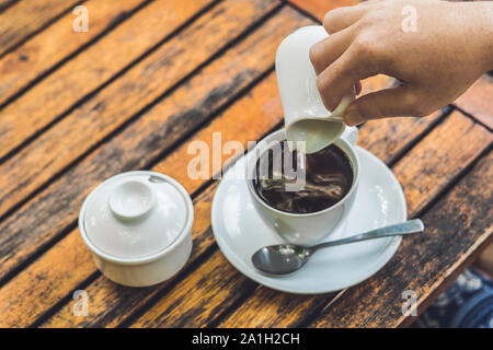 woman's hand pouring milk in white cup of coffee in outdoor cafe. Stock Photo
