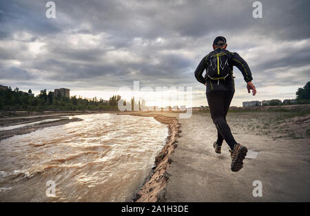 Man in black shirt with backpack running on the dirty trail near the river at sunset sky. Healthy lifestyle concept Stock Photo