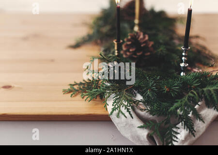 Stylish rustic christmas arrangement for festive dinner closeup. Fir branches with pine cones and vintage candlestick with black candles on linen clot Stock Photo