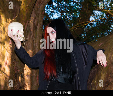 Dark and mysterious young female dressed in black goth costume talks to human skull  in medieval  church graveyard Stock Photo