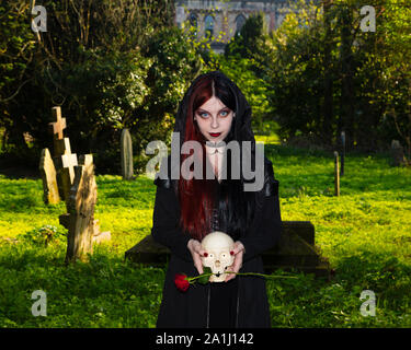 Dark and mysterious young female dressed in black goth costume talks to human skull  in medieval  church graveyard Stock Photo