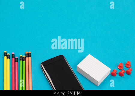 A half visible colorful pencils, black mobile phone, a stack of white scratch paper and a group of red pins are lying on a pale-blue surface isolated Stock Photo