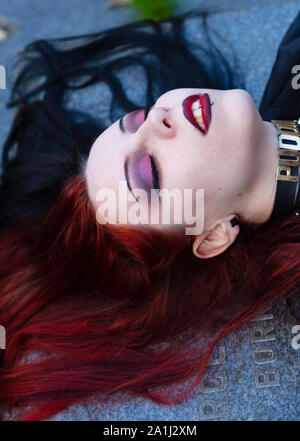 Dark  mysterious young female dressed in black goth  costume lying on old tombstone in medieval  church graveyard Stock Photo