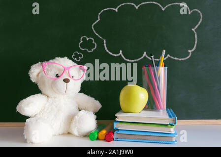 Cute teddy bear wearing glasses next to a pile of books and an apple in a classroom. Drawn thought bubble on a blackboard with empty copy space Stock Photo