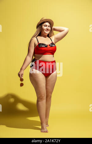Young caucasian plus size female model's preparing for beach resort on yellow background. Woman in red swimsuit, hat and sunglasses. Concept of summertime, party, body positive, equality and chill. Stock Photo