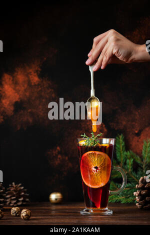 Mulled wine a warm drink made of red wine, citruses and spices in a glass on a wooden table. Female hand putting honey from a spoon into mulled wine Stock Photo