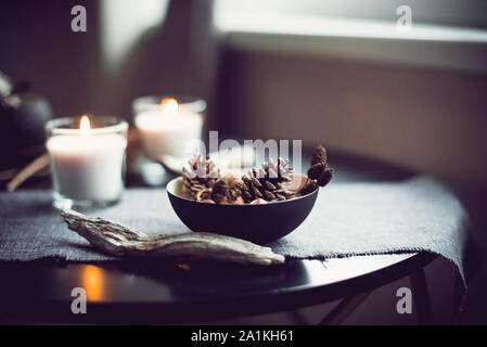 Burning candles and natural decor of cones, nuts in black bowl on the gray fabric napkin on the black table. Cozy atmosphere at home. Kinfolk Hygge Sl Stock Photo