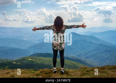 Girl traveling in mountains alone, standing with hands up achieving the top, welcomes a sun. Walking outdoors, woman hiker on mountain top. Wanderlust Stock Photo