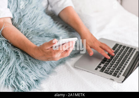 Female hands with a laptop and a phone are sitting on the bed. Modern business lady at home. Photo in light colors  Stock Photo