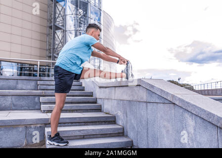 Male athlete, tanned man, summer in city, view from side, stretching muscles of legs knees ankle, warming up before jogging, fitness training workout Stock Photo